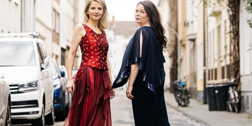 Zwei Frauen stehen auf einer städtischen Straße. Eine trägt ein rotes Kleid und schaur in die Kamera, die andere trägt blaue Kleidung, iläuft von der kamera weg und schaut dabei über die Schulter zurück. Am Straßenrand parken Autos.