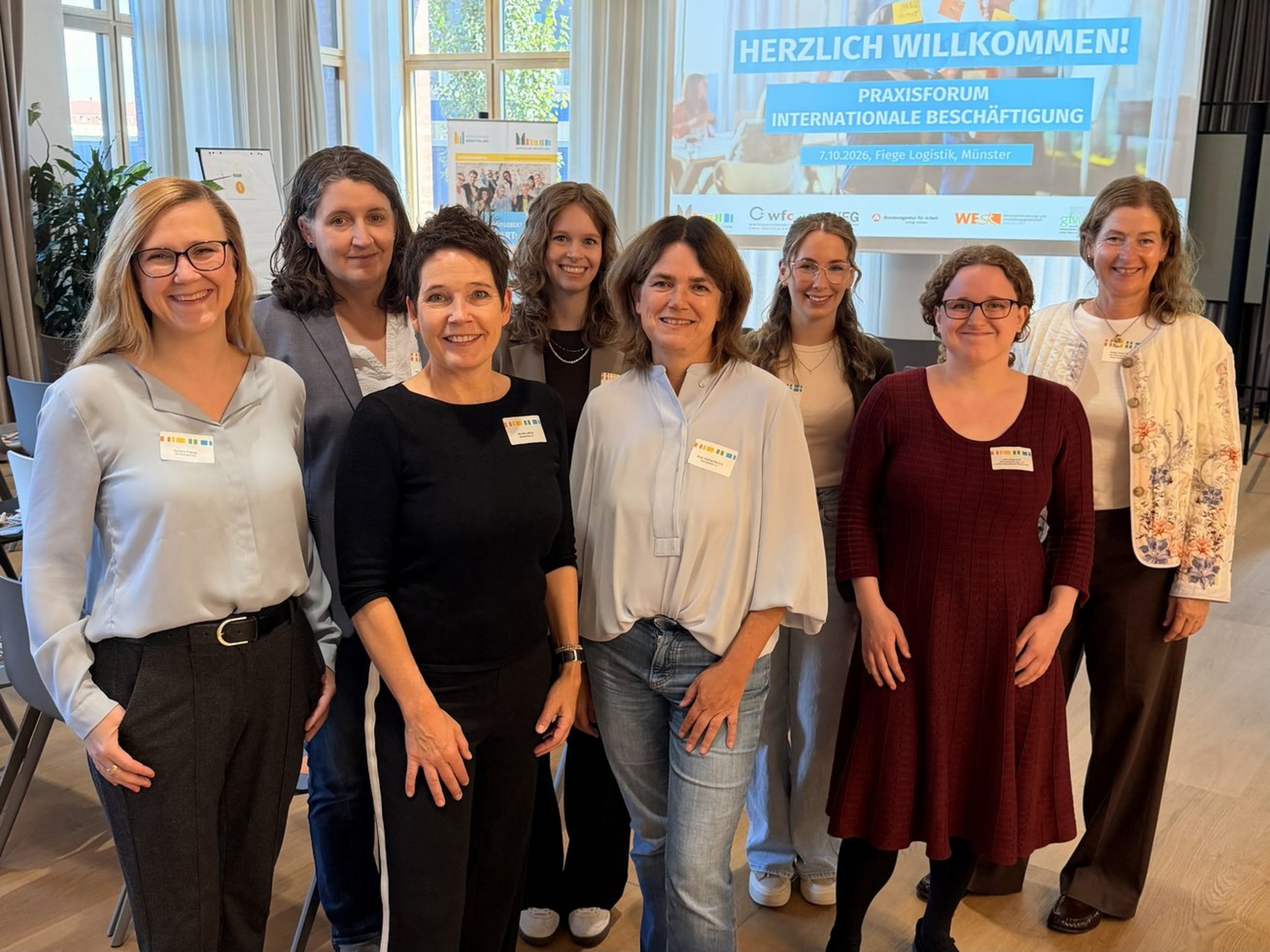 Acht Frauen stehen vor einer Leinwand auf die Herzlich Willkommen Praxisforum Internationale Beschäftigung projiziert wird.