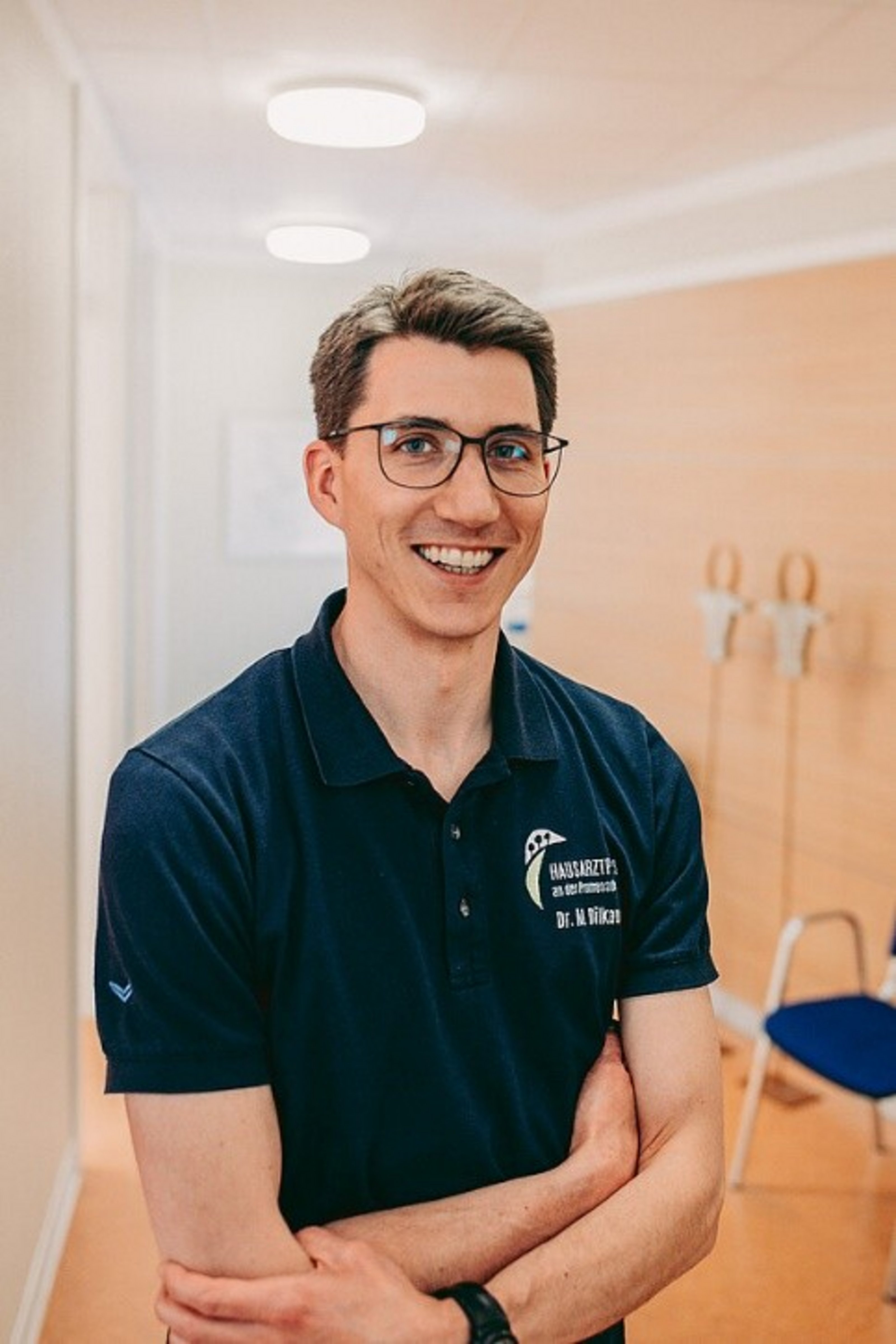 Hausarzt Dr. med. Matthias Dilkaute (Foto: Hannah Dürken) In Mann mit Brille und blauem Poloshirt steht mit verschränkten armen in einem hellen Raum mit braunem Fußboden.