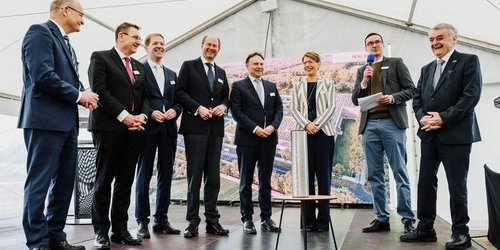 Mehrere Personen bei einer Grundsteinlegung auf einer Bühne, ein Redner mit Mikrofon spricht.