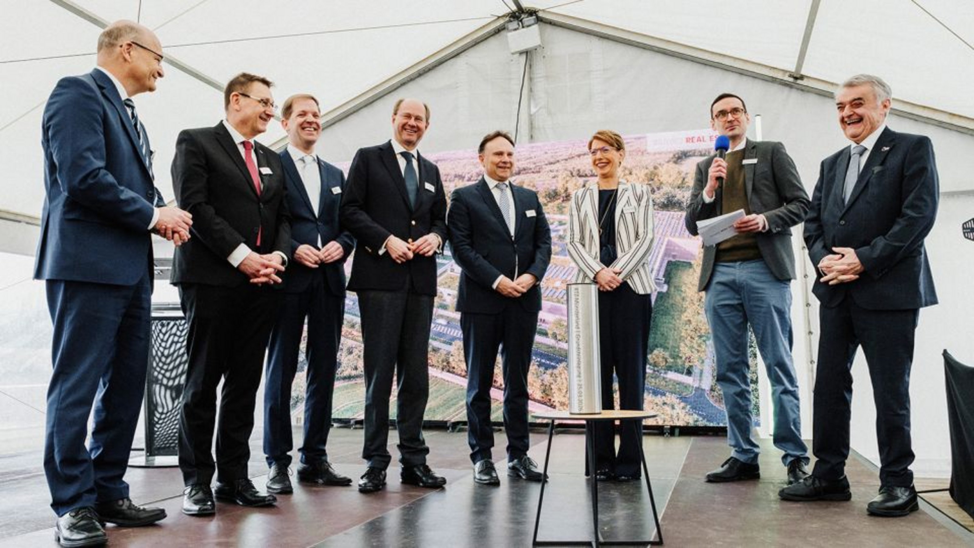 Mehrere Personen bei einer Grundsteinlegung auf einer Bühne, ein Redner mit Mikrofon spricht.