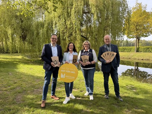 Vier Personen stehen nebeneinander auf einer Wiese an einem Fluss, der im Hintergrund fließt. Sie halten Papiertüten in der Hand, eine Person hält ein Schild mit der Aufschrift "Gut für mich. Gut für uns. Münsterland ... ist Klimaland."