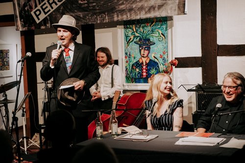 Eine Band sitzt und steht auf einer Bühne. Sie besteht aus vier Personen mit Instrumenten.