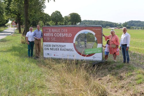 Am Straßenrand steht ein Banner mit der Aufschrift "Hier baut der Kreis Coesfeld für Sie. Hier entsteht ein neuer Radweg". Links und rechts neben dem Banner stehen vier erwachsene und ein Kind.