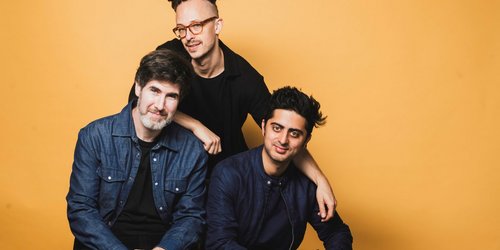 drei Männer in dunklen Oberteilen sitzen und hocken vor einem gelben Hintergrund.