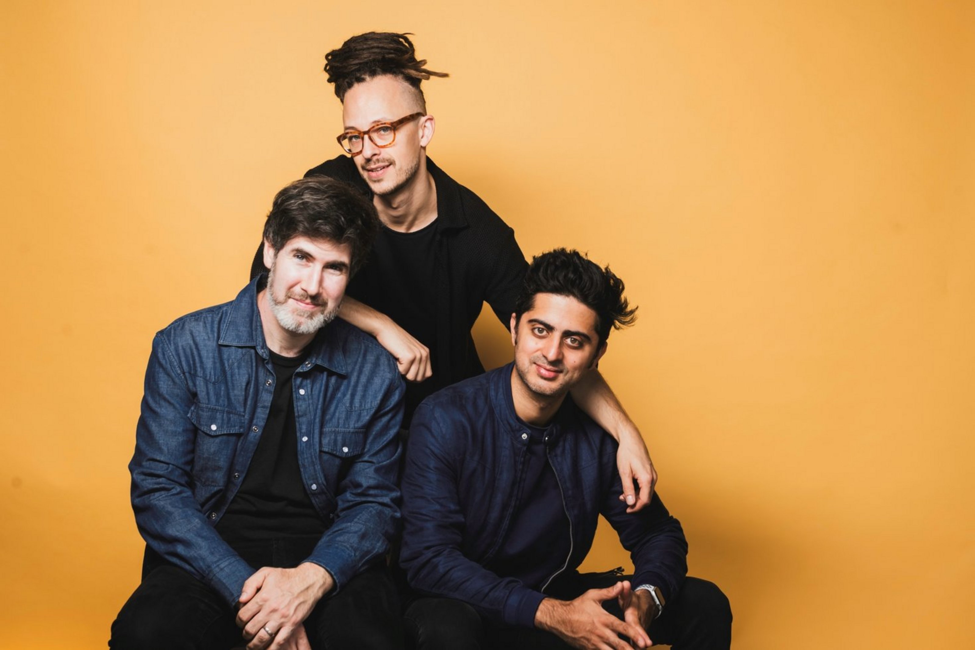 drei Männer in dunklen Oberteilen sitzen und hocken vor einem gelben Hintergrund.