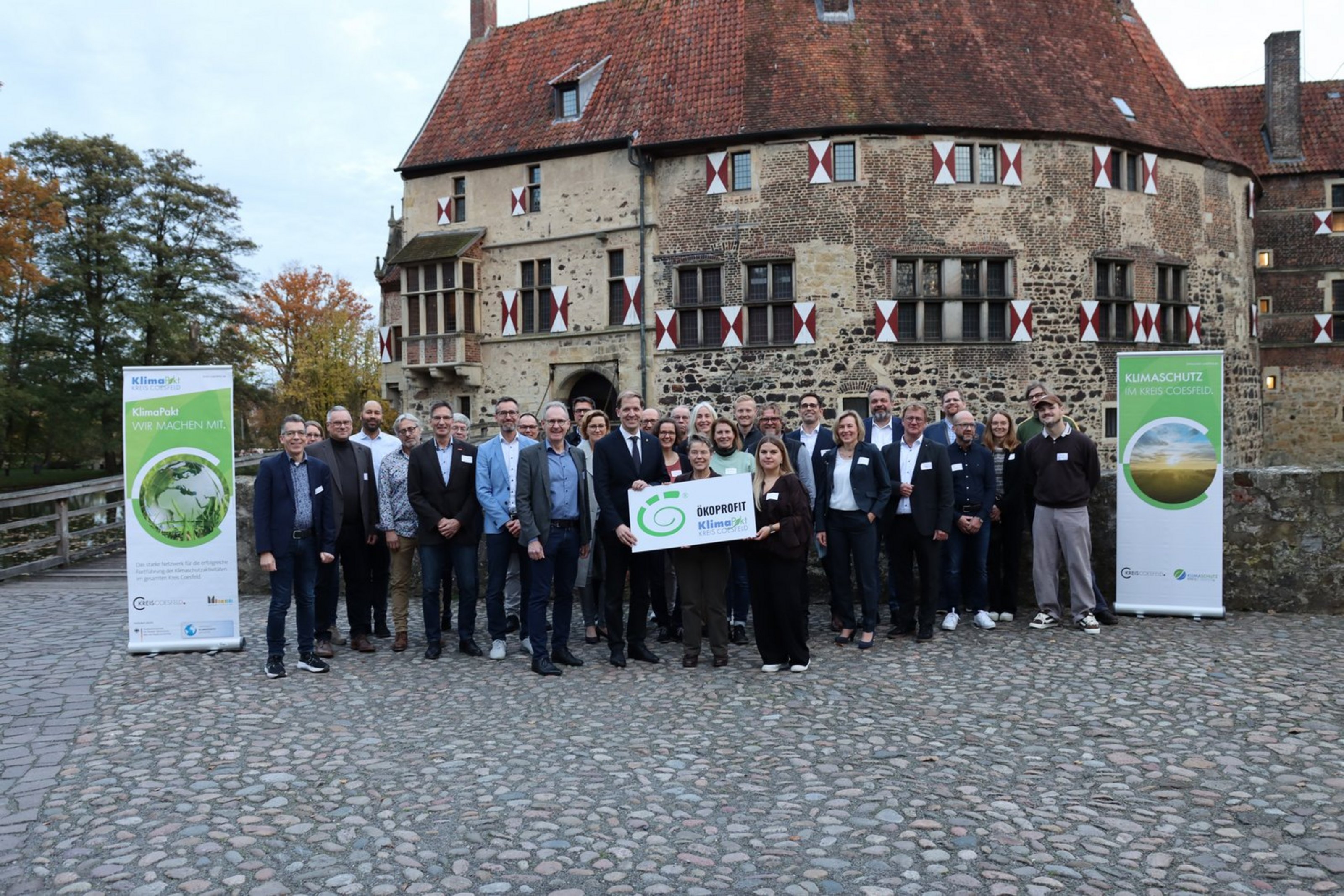 Eine Gruppe steht vor einer Burg und hält ein Schild mit der Aufschrift "Ökoprofit"."