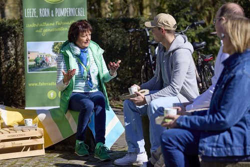 Eine Frau sitzt auf einem Hocker vor einem Aufsteller mit der Aufschrift "Leeze, Korn und Pumpernickel". Sie blickt drei Personen an, die ebenfalls auf Hockern sitzen und eine Tasse in der Hand halten.