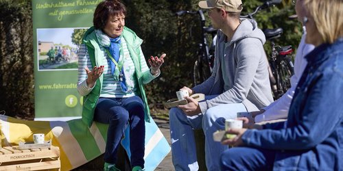 Eine Frau sitzt auf einem Hocker vor einem Aufsteller mit der Aufschrift "Leeze, Korn und Pumpernickel". Sie blickt drei Personen an, die ebenfalls auf Hockern sitzen und eine Tasse in der Hand halten.