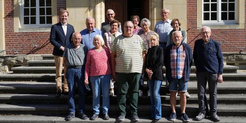 Dreizehn Seniorinnen und Senioren und ein Mann mittleren Alters stehen zusammen auf einer Eingangstreppe vor einem großen Haus.