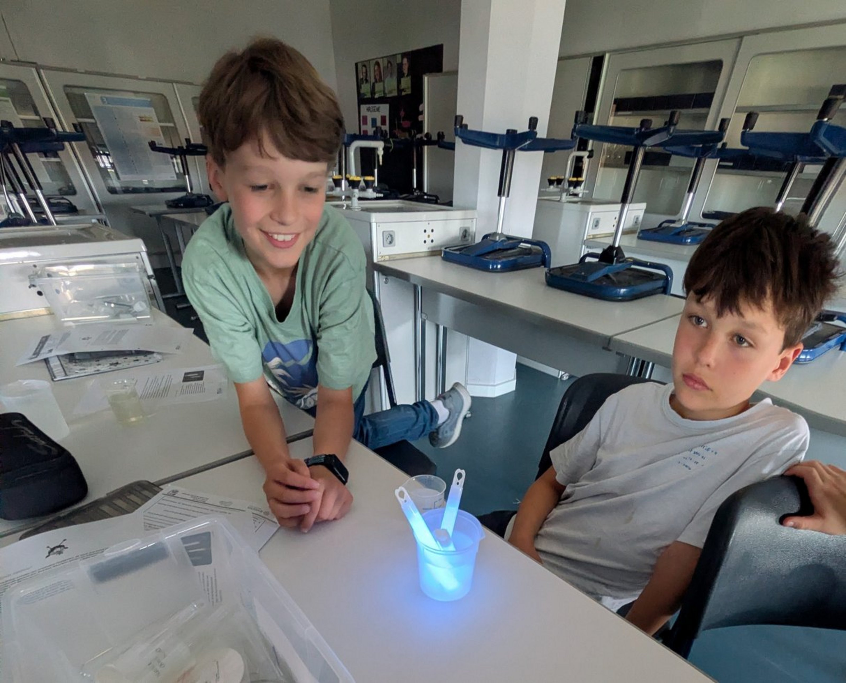 Zwei Kinder sitzen an einem Tisch. Vor ihnen steht ein Glasgefäß mit Wasser, in dem ein blaues Knicklicht platziert ist.