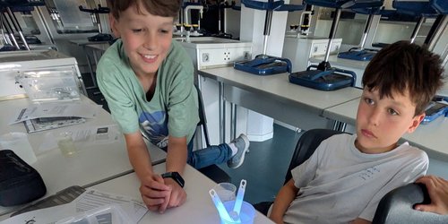 Zwei Kinder sitzen an einem Tisch. Vor ihnen steht ein Glasgefäß mit Wasser, in dem ein blaues Knicklicht platziert ist.