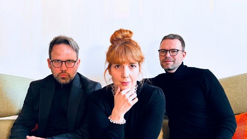 Drei Personen sitzen nebeneinander. Sie tragen alle dunkle Oberteile . Die Frau in der Mitte hat die Hand ans Kinn gelegt. Die Männer tragen eine Brille.