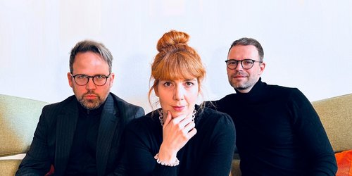 Drei Personen sitzen nebeneinander. Sie tragen alle dunkle Oberteile . Die Frau in der Mitte hat die Hand ans Kinn gelegt. Die Männer tragen eine Brille.