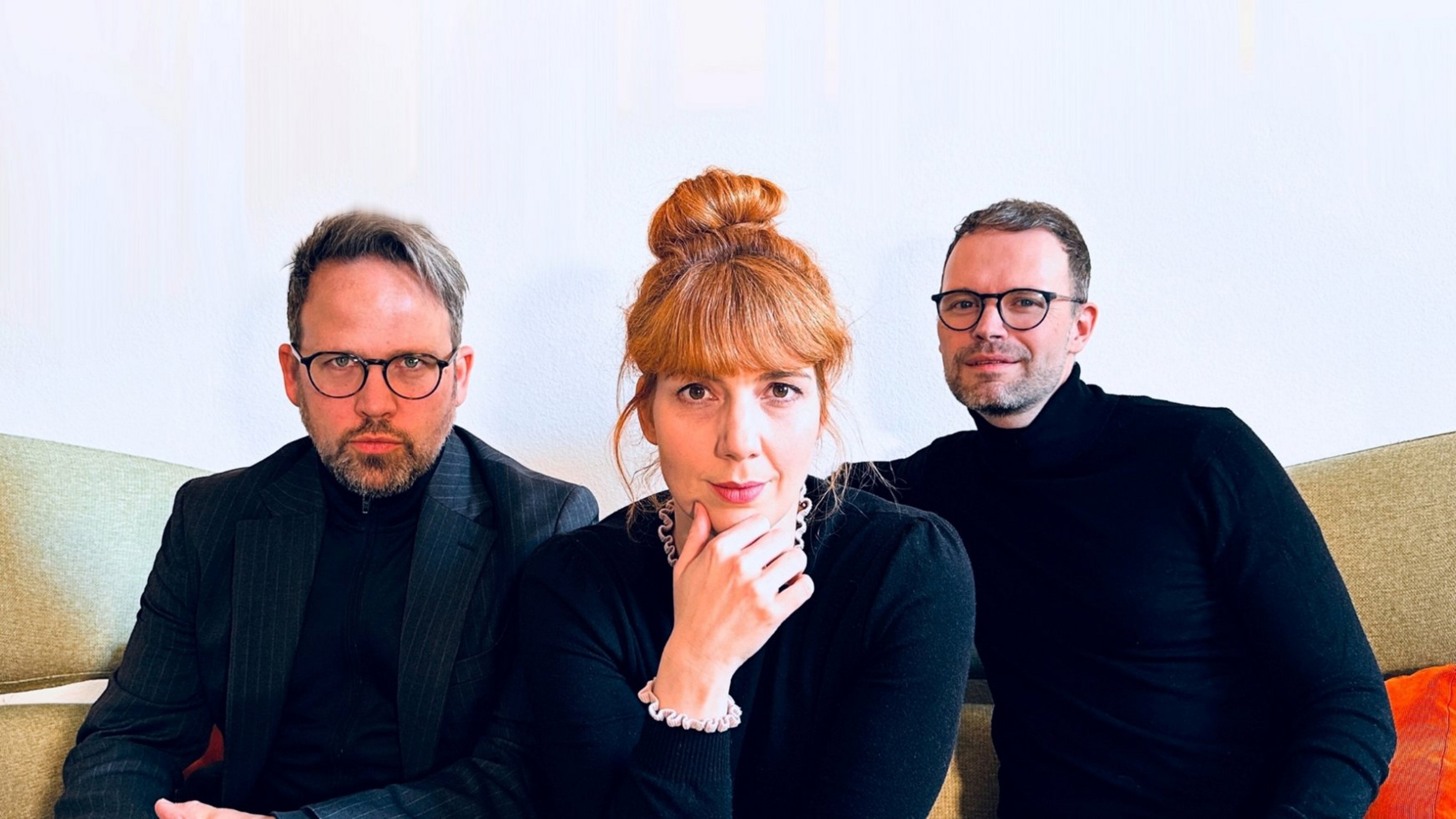 Drei Personen sitzen nebeneinander. Sie tragen alle dunkle Oberteile . Die Frau in der Mitte hat die Hand ans Kinn gelegt. Die Männer tragen eine Brille.