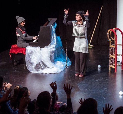 Auf einer Theaterbühne findet eine musikalische Aufführung für Kinder statt. Links sitzt eine Frau in winterlicher Kleidung mit Mütze am Klavier, das mit transparentem, weiß-blauem Stoff dekoriert ist. Rechts steht eine weitere Frau, ebenfalls winterlich gekleidet, und spricht oder singt mit erhobenen Händen zu einem jungen Publikum, das im Vordergrund sichtbar ist und begeistert die Hände hebt. Im Hintergrund sind Requisiten wie ein Schlitten, ein Besen und Schneebälle zu sehen. Die Szene vermittelt eine winterliche Atmosphäre und eine interaktive, kindgerechte Vorstellung.