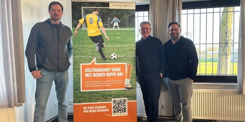 Drei Männer stehen neben einem Banner, auf dem "Zieltraining? Fang mit deiner Kippe an!" steht und einen Fußballspieler zeigt, der einen Ball in Richtung eines Fußballtores mit Torwart schießt.