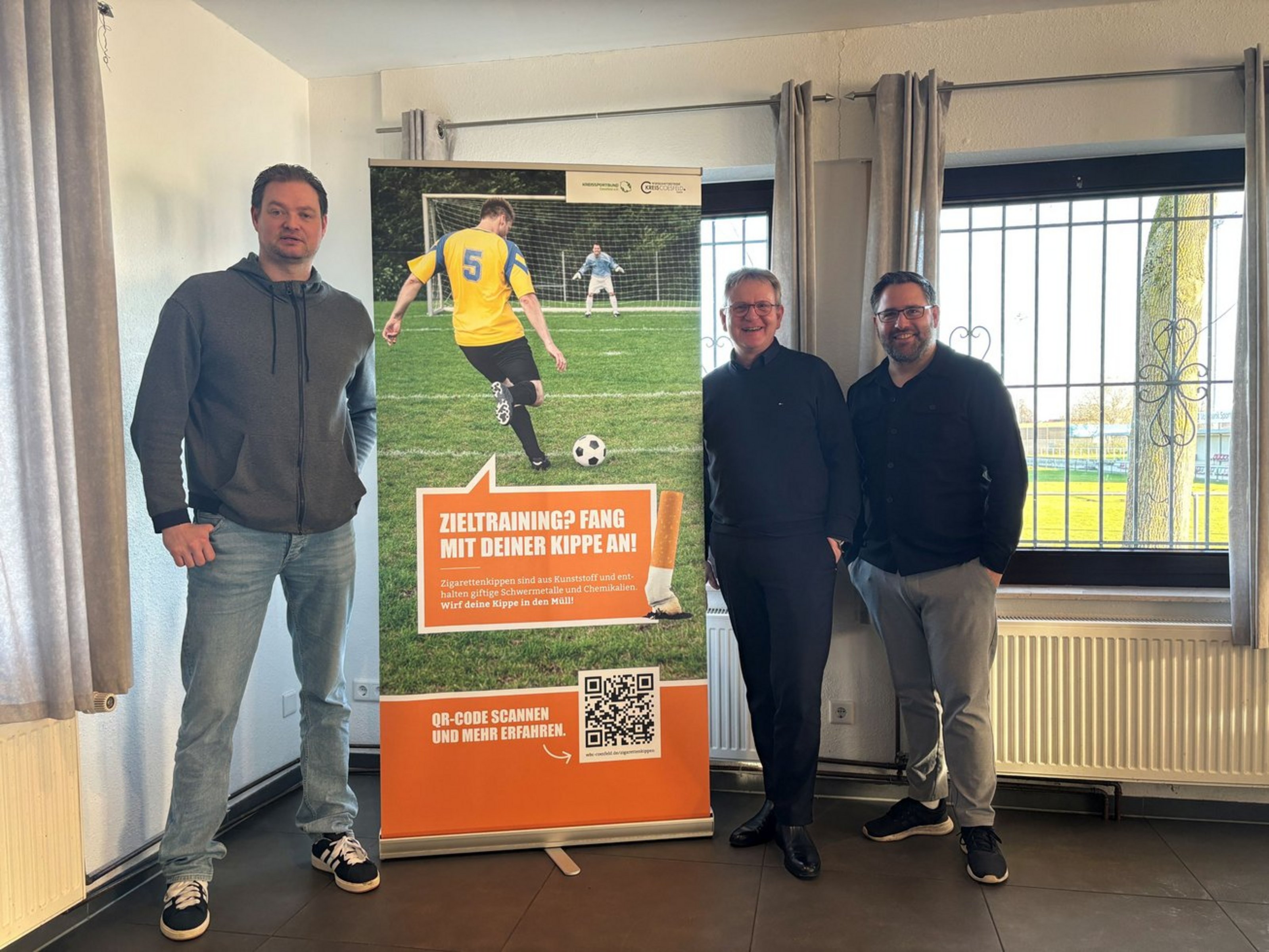 Drei Männer stehen neben einem Banner, auf dem "Zieltraining? Fang mit deiner Kippe an!" steht und einen Fußballspieler zeigt, der einen Ball in Richtung eines Fußballtores mit Torwart schießt.