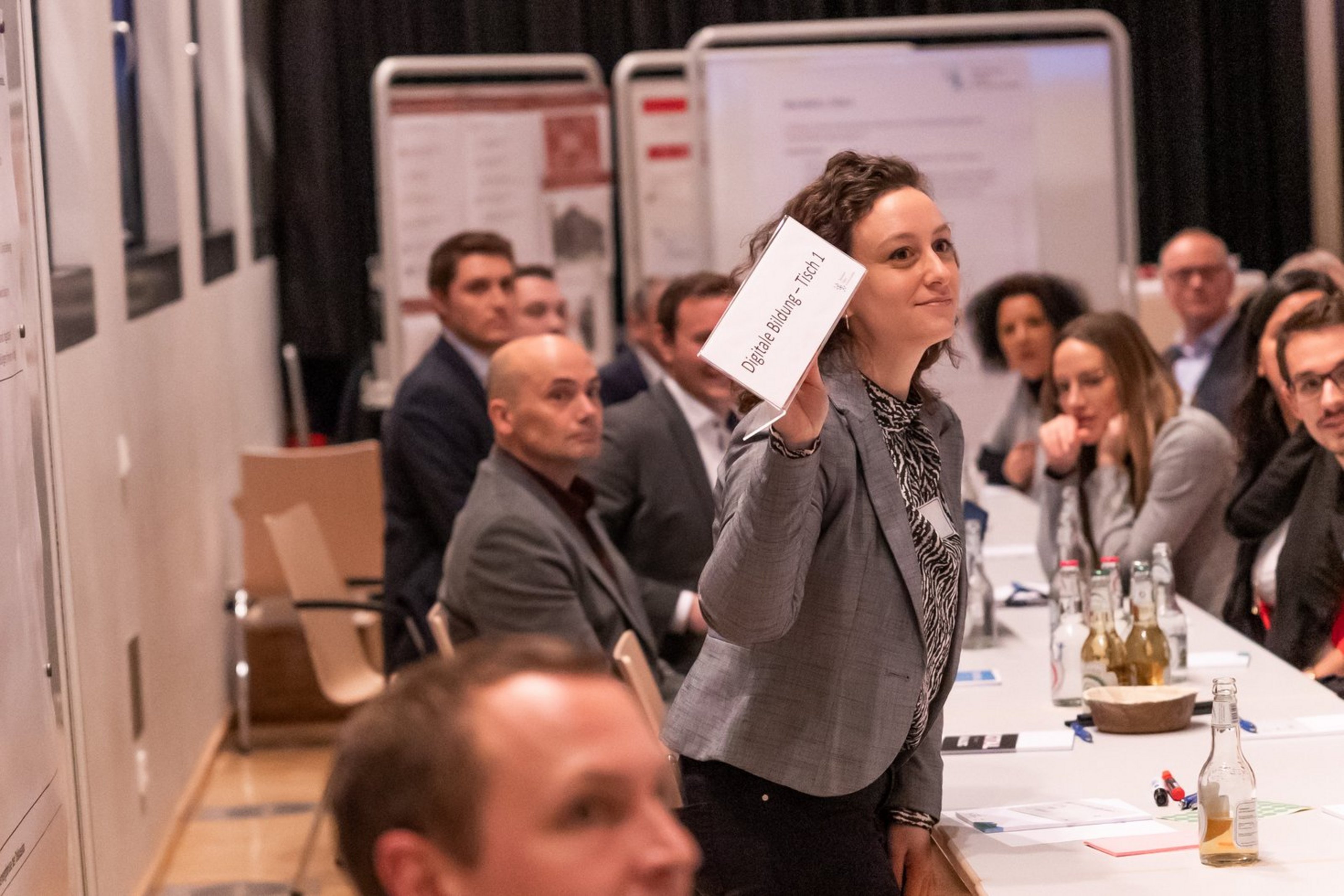 Frau hält ein Blatt Papier vor einer Gruppe sitzender Menschen in einem Konferenzraum