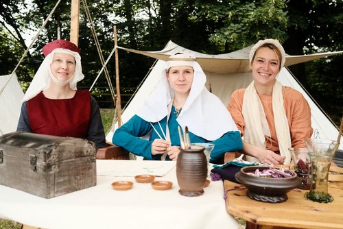 Drei Frauen in historischer mittelalterlicher Kleidung sitzen vor Zelten im Freien. Sie tragen Kopfbedeckungen und sind in traditionelle Gewänder gehüllt. Vor ihnen auf den Tischen stehen verschiedene Gegenstände wie ein hölzernes Kästchen, Ton- und Glasgefäße sowie Schalen mit Kräutern oder Garn.