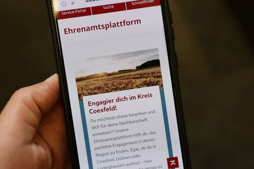 Eine Handybildschirm ist zu sehen, eine Hand hält das Handy fest. Auf dem Bildschirm steht " Ehrenamtsplattform" sowie "Engagier dich im Kreis Coesfeld!".