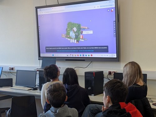 Fünf Kinder schauen auf einen Bildschirm, der über einem Tisch mit Computern hängt. 