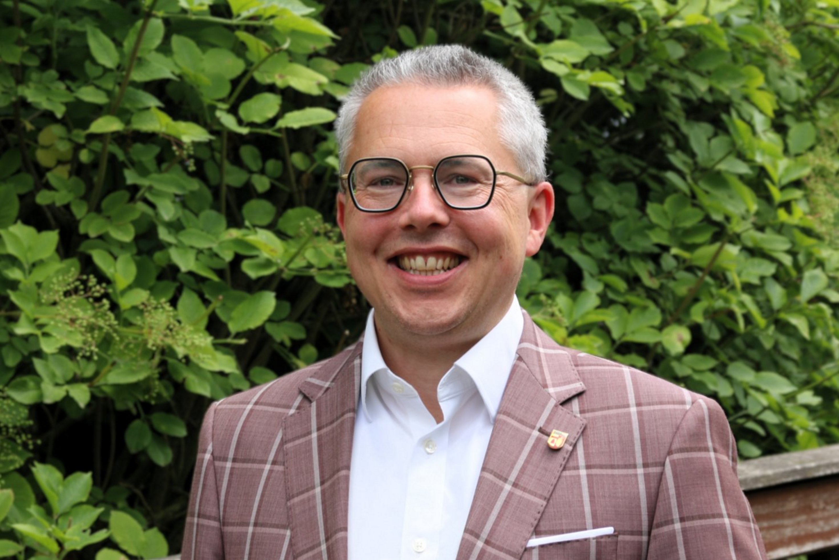 Ein Mann mit kurzen Haaren und Brille trägt eine karierte Anzugjacke und lächelt.