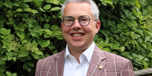 Ein Mann mit kurzen Haaren und Brille trägt eine karierte Anzugjacke und lächelt.
