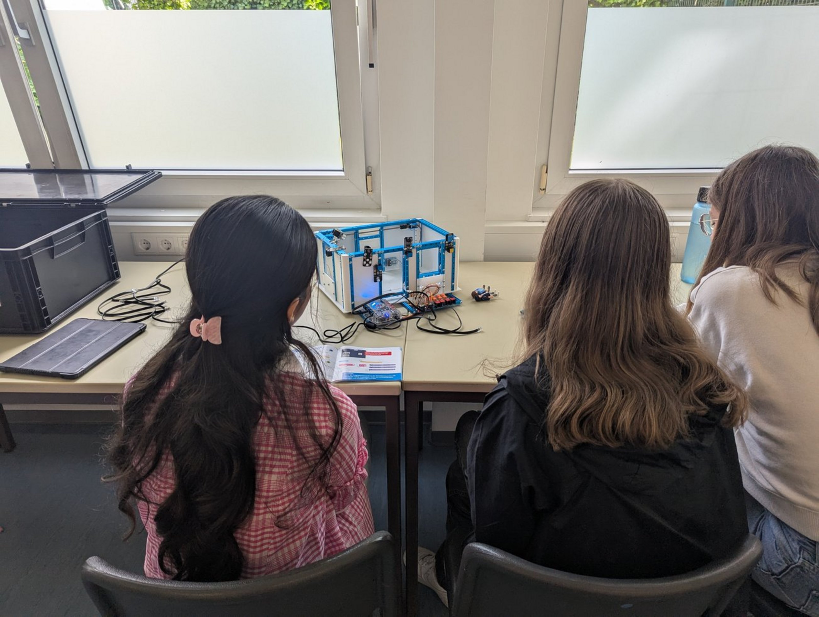 Drei Schülerinnen steuern ein Smarthome-Modell am Tisch mit Tablet und Baukasten.