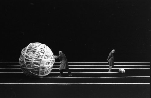 Man sieht ein künsterlisches Foto von einem Mann, der ein überdimensionalen Wollball vor sich her schiebt.