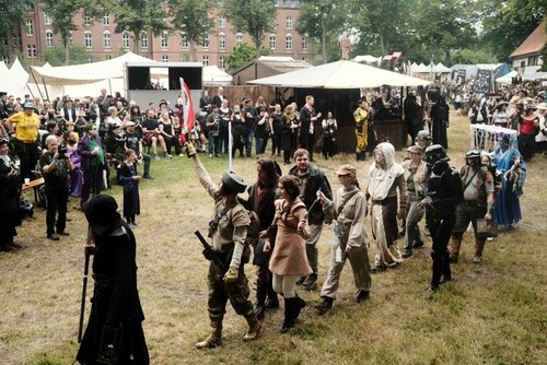 Über eine Wiese mit vielen schaulustigen läuft eine kostümierte Gruppe nacheinander in einer langen Schlange. Im Hintergrund isnd weiße Zelte zu sehen. Die Gruppe wir von einer Person in einer schwarzen Kutte angeführt.