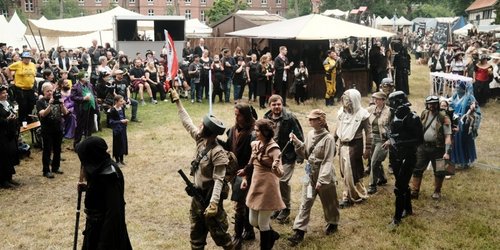 Über eine Wiese mit vielen schaulustigen läuft eine kostümierte Gruppe nacheinander in einer langen Schlange. Im Hintergrund isnd weiße Zelte zu sehen. Die Gruppe wir von einer Person in einer schwarzen Kutte angeführt.