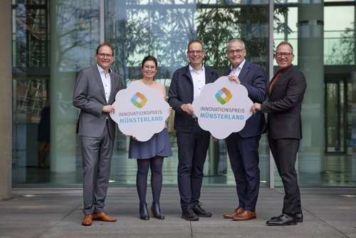 Fünf Personen stehen nebeneinander vor einer Glasfront . Sie halten zwei Schilder mit der Aufschrift "Innovationspreis Münsterland".