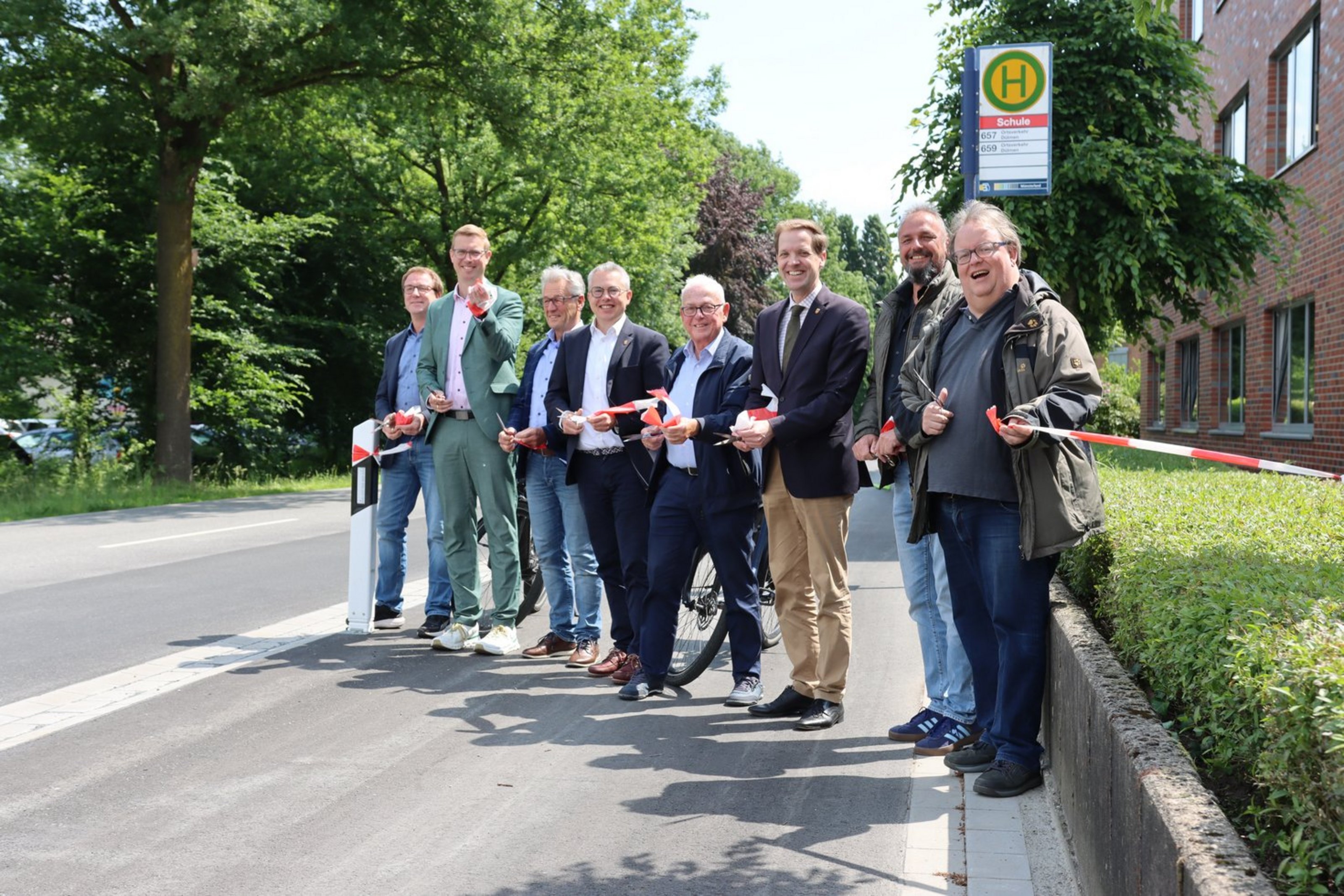 Freigabe des Radwegs entlang der Kreisstraße K27 (Foto: Kreis Coesfeld). Acht Männer stehen nebeneinander auf einem Radweg neben einer Straßen. Sie halten rot-weißes Absperrband und Scheren in den Händen. Hinter der Gruppe ist ein Bushaltestellenschild mit der Aufschrift "Schule" zu sehen. Rechts befindet sich ein rot geklinkertes Gebäude, entlang der Straße stehen Bäume.