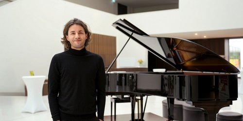 Ein Mann steht mit Händen in den Hosentaschen vor einem Klavier in einem hellen Saal.