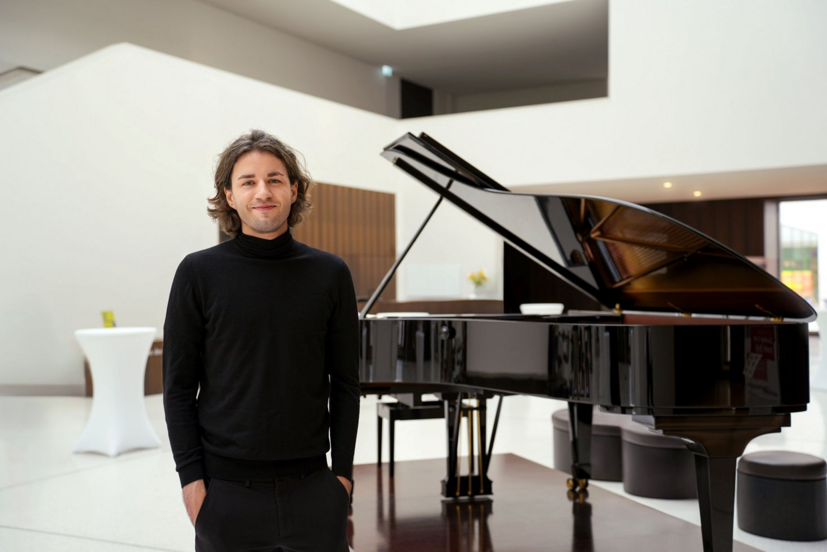 Ein Mann steht mit Händen in den Hosentaschen vor einem Klavier in einem hellen Saal.