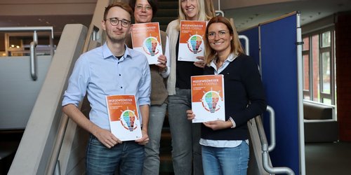 Auf einer Treppe stehen vier Personen, jeweils auf zwei Treppenstufen und halten eine orange Broschüre in den Händen. Sie lächeln in die Kamera.