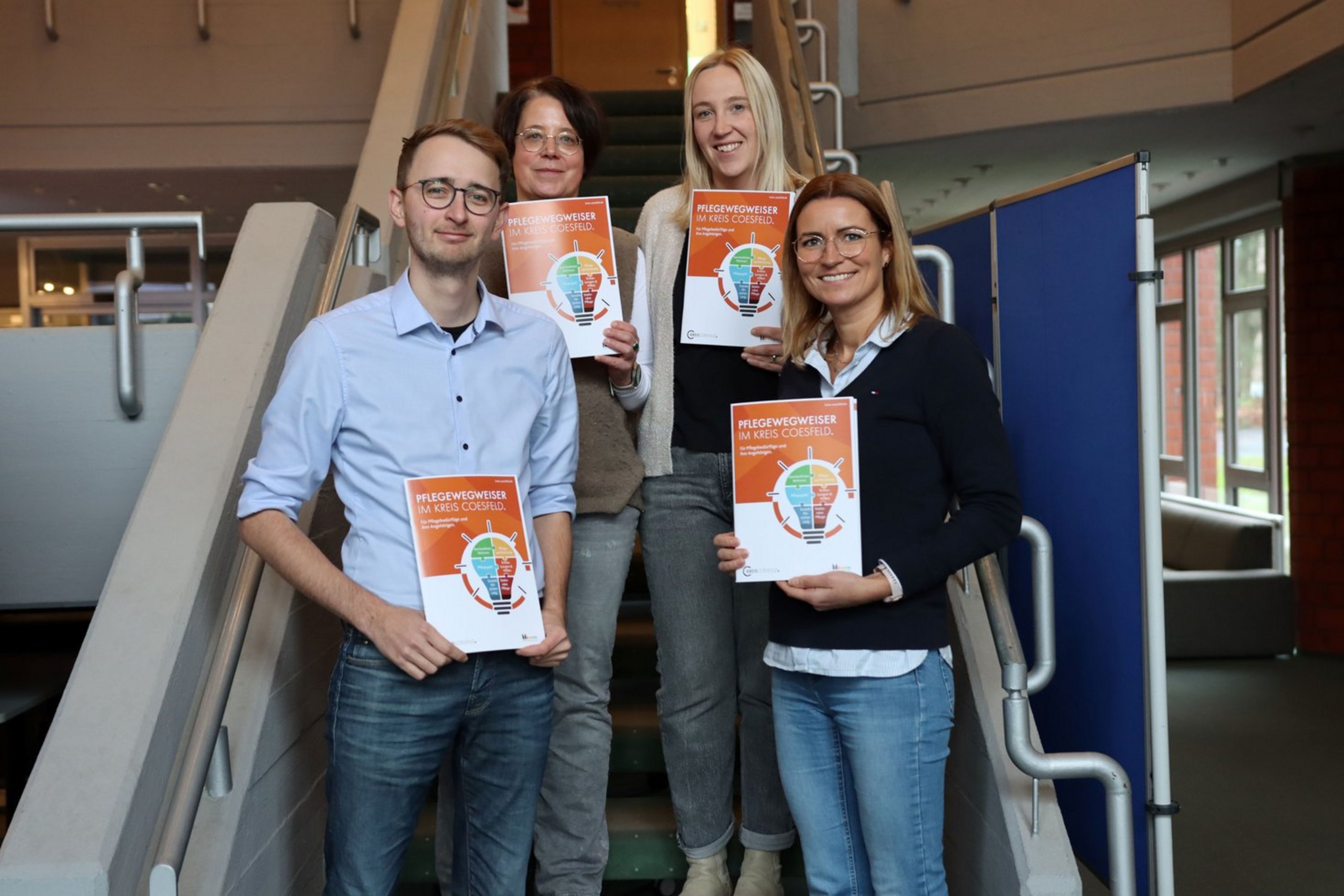 Auf einer Treppe stehen vier Personen, jeweils auf zwei Treppenstufen und halten eine orange Broschüre in den Händen. Sie lächeln in die Kamera.