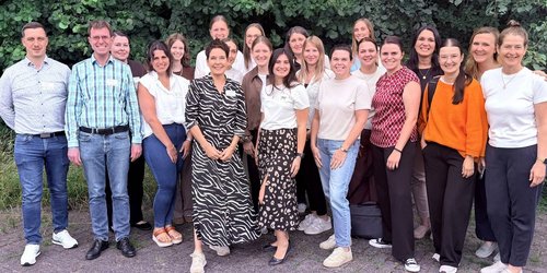 Einundzwanzig Männer und Frauen stehen in einer Gruppe zusammen vor einem Waldstück und schauen fröhlich Richtung Kamera.