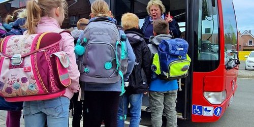 Kinder stehen in einer Schlange vor einem Bus mit geöffnete Tür. Auf der Anzeige des Busses steht Busschule.