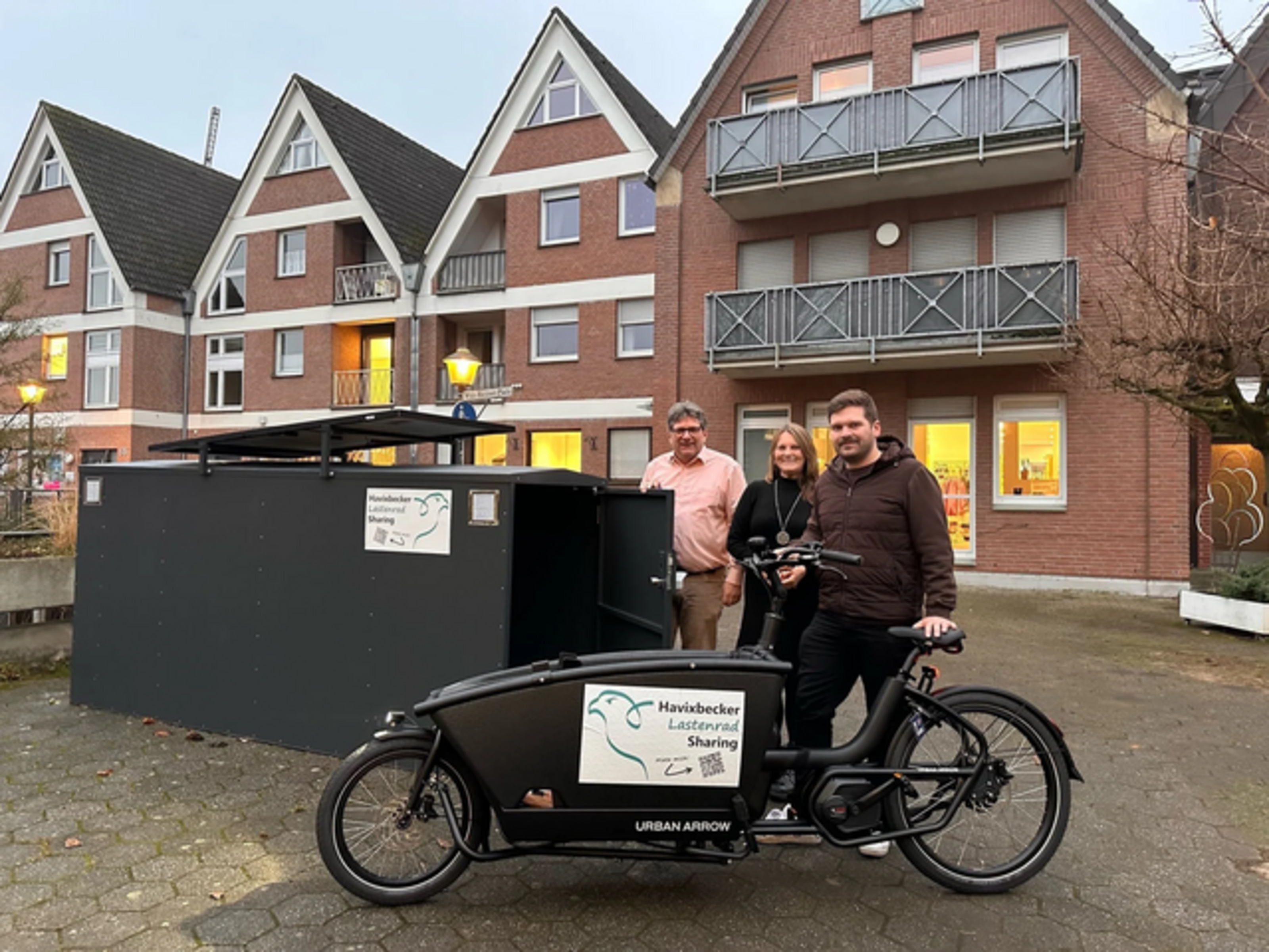 Drei Personen stehen hinter einem schwarzen Lastenfahrrad mit Aufkleber "Havixbecker Lastenrad Sharing" vor einem Wohnhaus mit Giebel und Balkonen