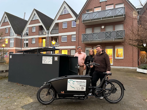 Drei Personen stehen hinter einem schwarzen Lastenfahrrad mit Aufkleber "Havixbecker Lastenrad Sharing" vor einem Wohnhaus mit Giebel und Balkonen