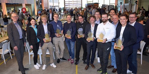 Eine Gruppe aus 14 Personen, davon 12 Männer und eine Frau, stehen in einer gutbesuchten Messehalle. Einige von Ihnen halten Preise in der Hand.