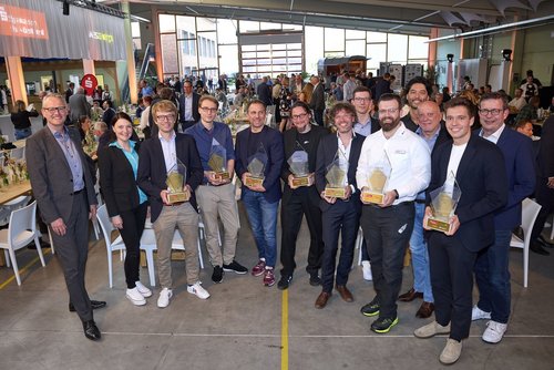 Eine Gruppe aus 14 Personen, davon 12 Männer und eine Frau, stehen in einer gutbesuchten Messehalle. Einige von Ihnen halten Preise in der Hand.