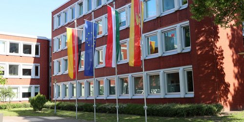 Vor einem mehrstöckigem Gebäude aus rotem Stein stehen vier Fahnenmasten, an denen die Flaggen der Bundesrepublik Deutschland, Europas, Nordrhein-Westfalens und des Kreises Coesfeld hängen.