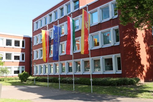 Vor einem mehrstöckigem Gebäude aus rotem Stein stehen vier Fahnenmasten, an denen die Flaggen der Bundesrepublik Deutschland, Europas, Nordrhein-Westfalens und des Kreises Coesfeld hängen.
