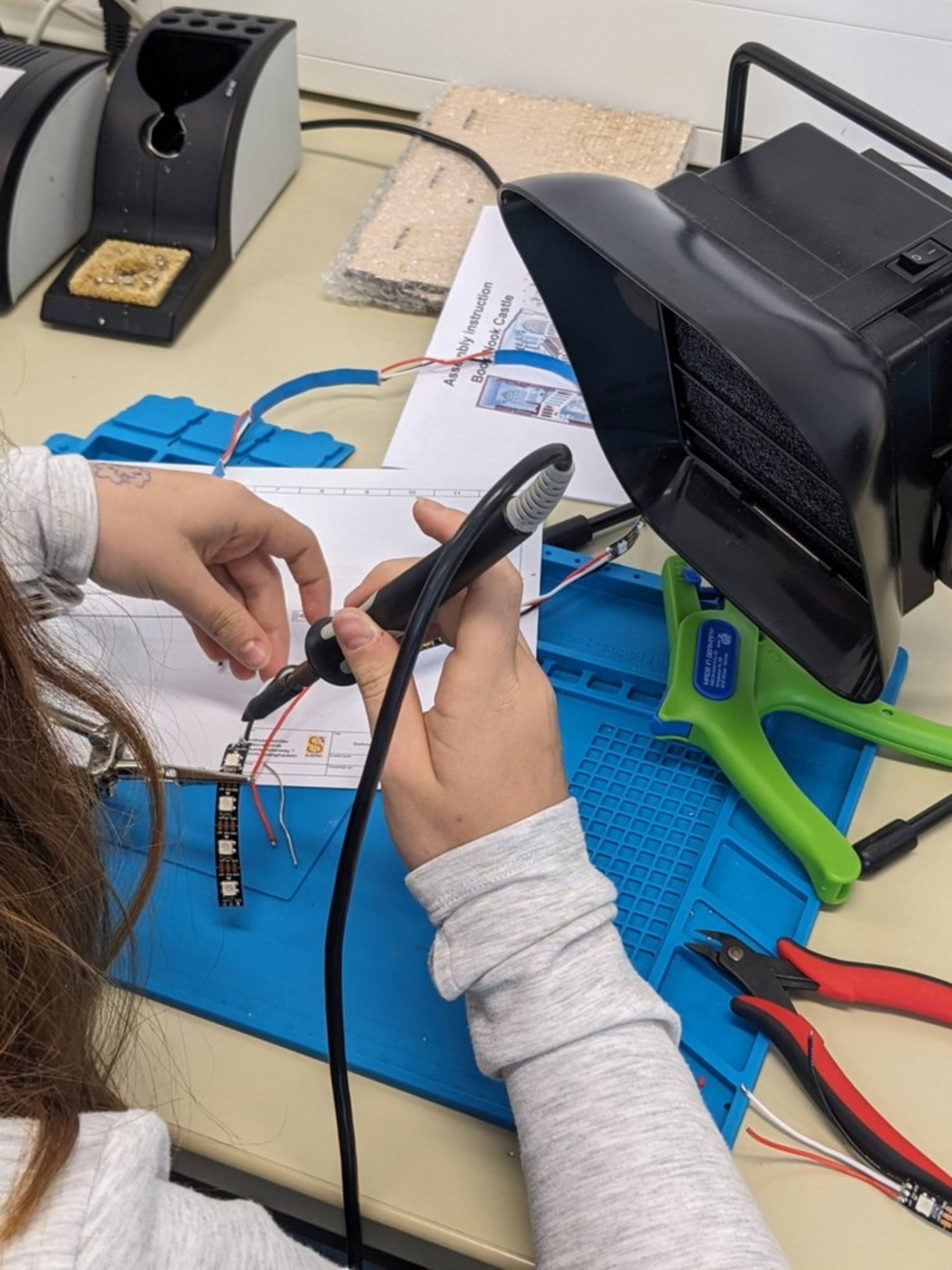 Kinderhände arbeiten mit Lötkolben und Elektronikbauteilen an einem Tisch.