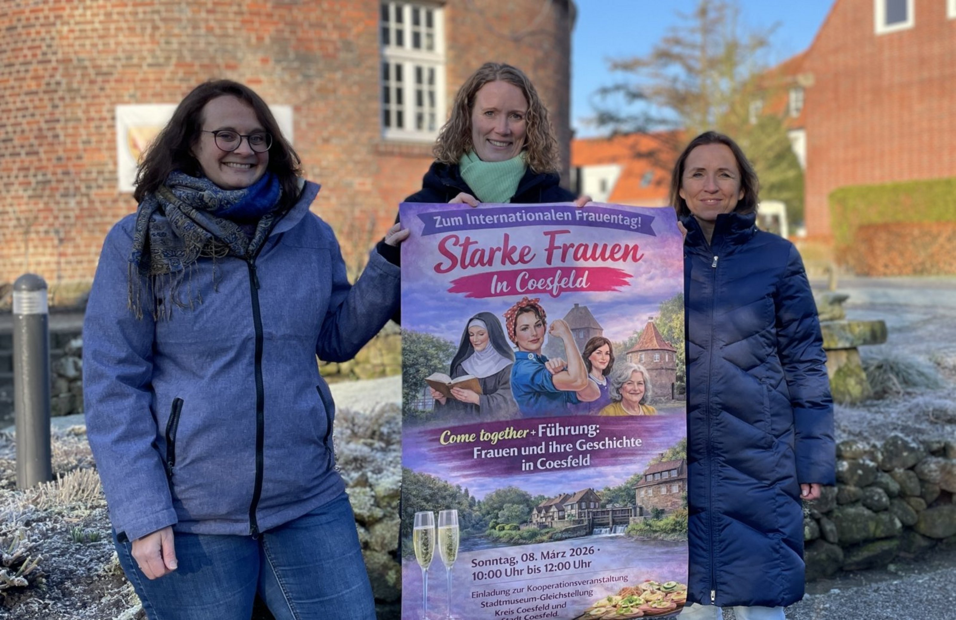 Drei Frauen stehen draußen vor einer Hecke und einem Backsteingebäude im Hintergrund und halten gemeinsam ein Plakat, auf dem "Starke Frauen" steht.