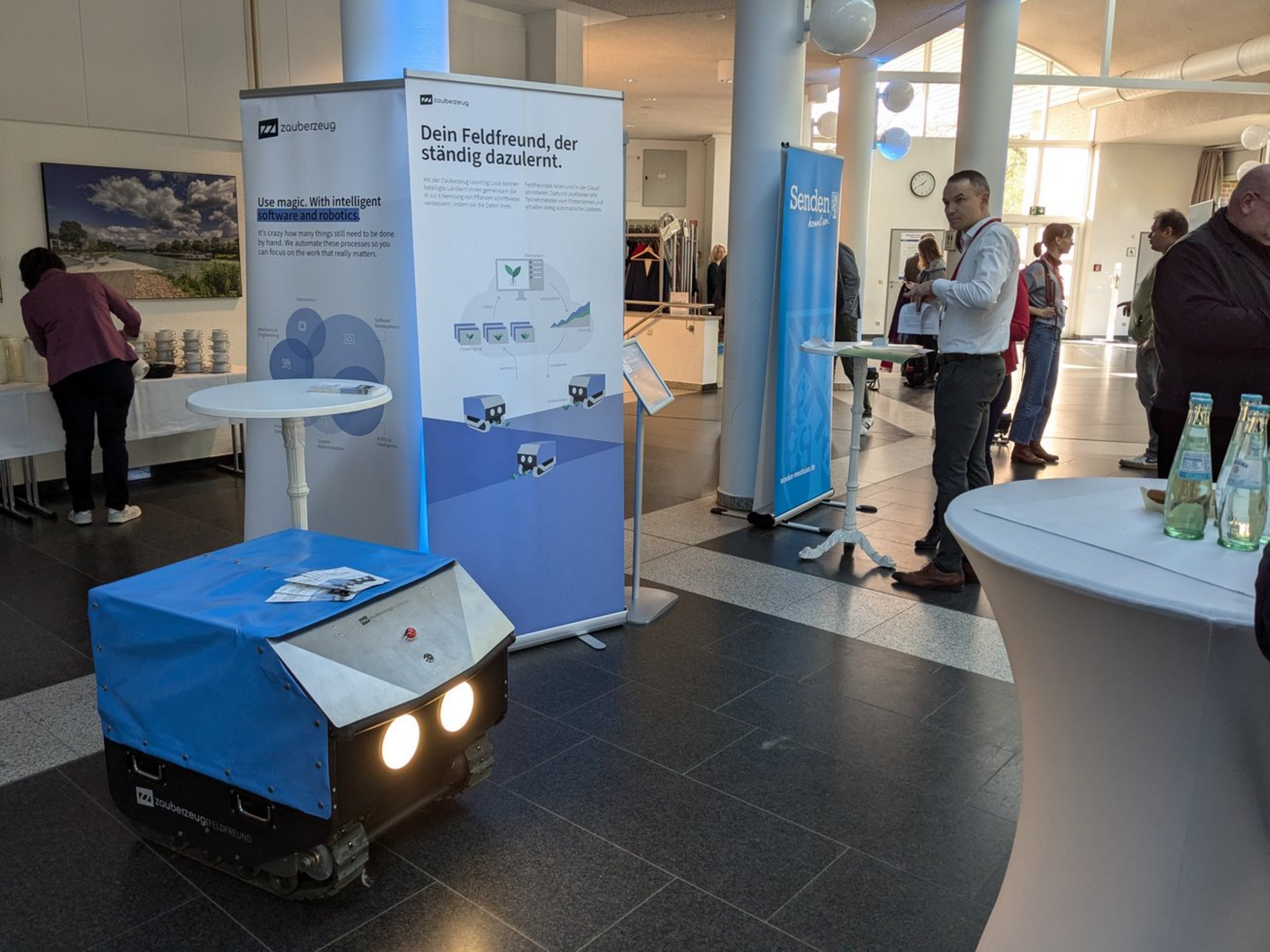 Kleiner mobiler Roboter mit zwei leuchtenden Scheinwerfern und blauem Überzug steht vor Informationsrollups auf einer Veranstaltung mit mehreren Personen und Stehtischen.