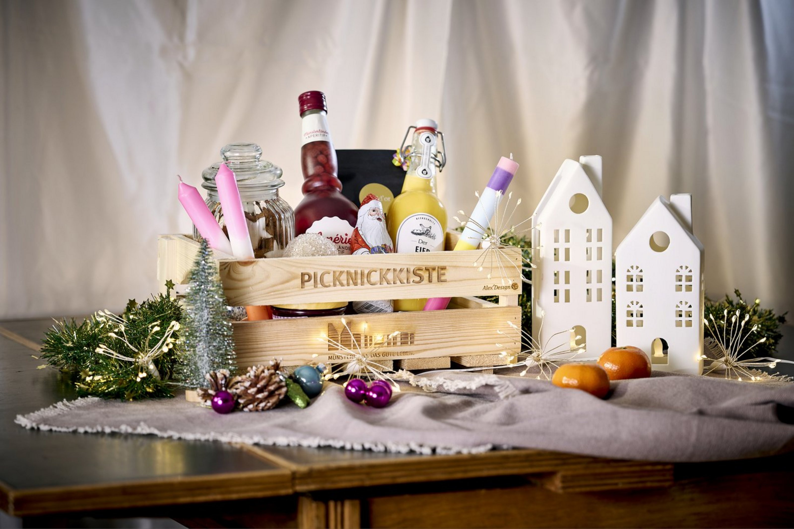 Auf einem Tisch steht eine Holzkiste. Daneben stehen verschiedene Dekoartikel, die mittels der Münsteraner Giebelhäuser und weihnachtlicher Dekoration ein festliche Stimmung schaffen. In der Kiste sind verschiedene Flaschen und Verpackungen mit Spezialitäten aus dem Münsterland zu sehen.